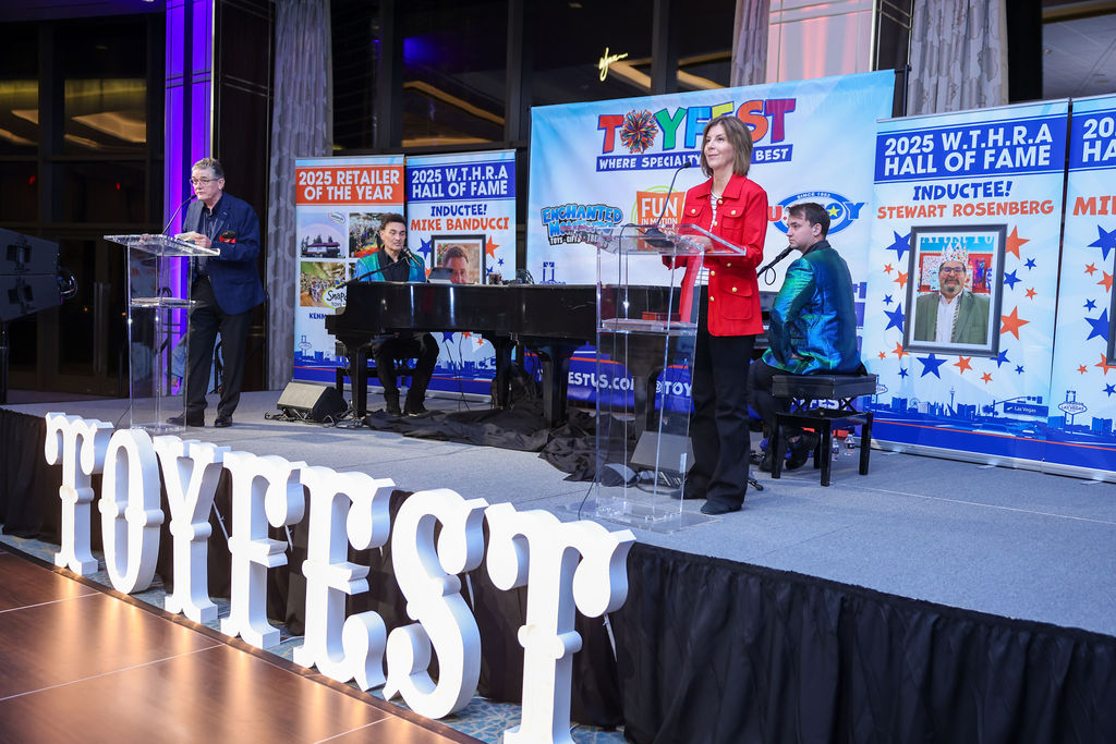 A stage setup at Toyfest featuring a man speaking at a podium, a woman in a red blazer speaking from another podium, and a young man playing piano in the background. Banners displaying event details and inductees are visible behind them.