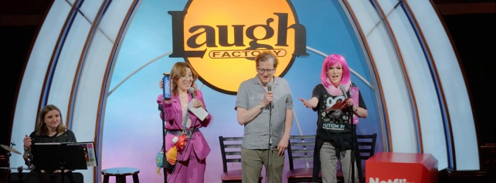 A stand-up comedy performance at the Laugh Factory with three performers on stage: a man in a button-up shirt holding a microphone, a woman in a pink suit holding a drink, and another woman with pink hair gesturing expressively. In the background, there is a sign for the Laugh Factory and a Netflix logo on a stool.