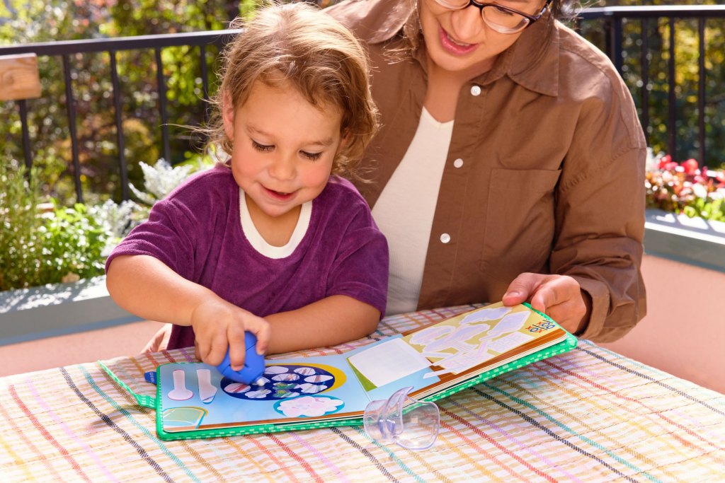 A young child and an adult are engaged in an activity together, exploring a colorful book on a table outdoors. The child uses a blue tool to interact with the book, while the adult smiles and observes.