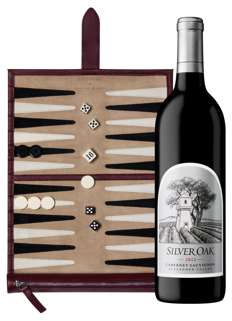 A bottle of Silver Oak 2022 Cabernet Sauvignon stands next to a stylish backgammon board featuring dice and game pieces, presented in a burgundy and beige color scheme.